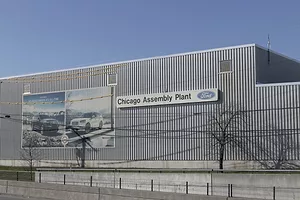 EXTERIOR OF A FORD ASSEMBLY PLANT. NEXT TO A LARGE IMAGE OF FORD VEHICLES IS A SIGN THAT READS "CHICAGO ASSEMBLY PLANT" FOLLOWED BY THE FORD LOGO.