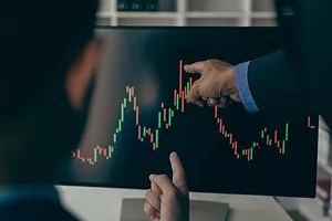 TWO PEOPLE SITTING AT A COMPUTER WITH MARKET STATISTICS ON IT. ONE OF THE PEOPLE IS POINTING AT THE COMPUTER MONITOR.