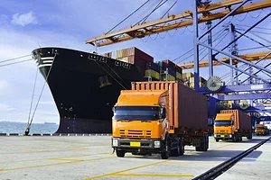 A FLEET OF THREE ORANGE TRUCKS DRIVE ON THE DOCK OF A PORT NEXT TO A LARGE CONTAINER SHIP.