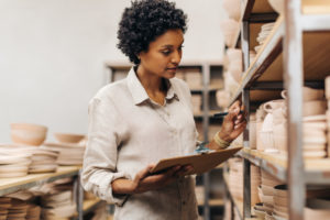 A WOMAN USES A PEN AND CLIPBOARD TO TRACK HER POTTERY INVENTORY.