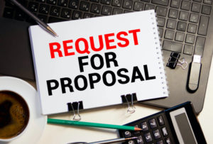 A NOTEPAD WITH THE PHRASE "REQUEST FOR PROPOSAL" ON IT SITS ON TOP OF A KEYBOARD SURROUNDED BY A CALCULATOR, A CUP OF COFFEE AND OTHER OFFICE SUPPLIES.