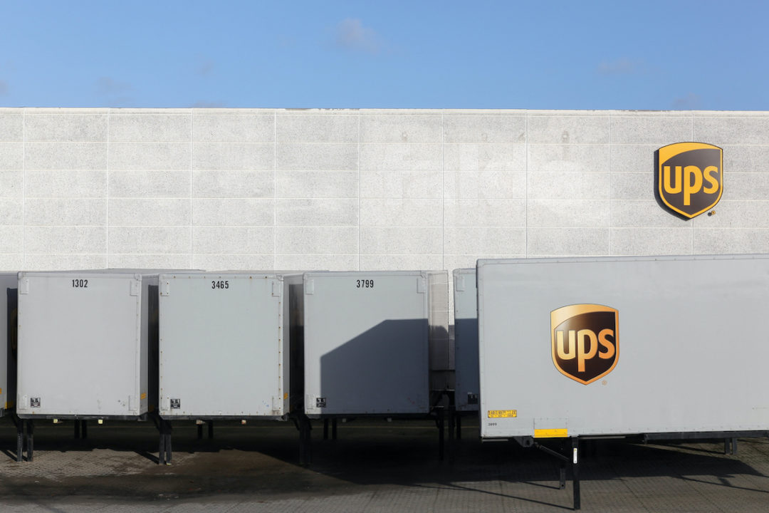 UPS LABELED TRACTOR TRAILERS ARE LINED UP OUTSIDE OF A WAREHOUSE WITH THE UPS LOGO ON IT.