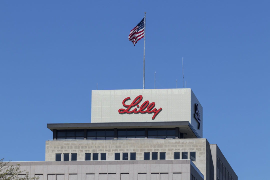 A BUILDING WITH THE ELI LILLY LOGO ON IT UNDER A CLEAR, BLUE SKY. AN AMERICAN FLAG WAVES ON A FLAGPOLE ABOVE THE LOGO.