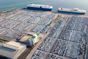 AERIAL SHOT OF THOUSANDS OF CARS AT A DOCK, WITH TWO SHIPS DOCKED NEARBY