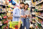 TWO PEOPLE IN A SUPERMARKET AISLE EXAMINE A GLASS JAR OF PRODUCT HELD BY ONE OF THEM