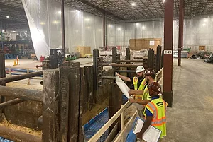 WORKERS IN HI-VIS VESTS DISCUSS A SHEET OF PLANS IN THE MIDST OF CONSTRUCTION IN A WAREHOUSE SPACE.