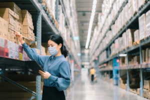 A MASKED WOMAN USING A TABLET CAN BE SEEN CHECKING WAREHOUSE INVENTORY.