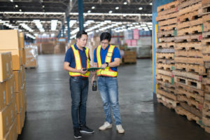 TWO WAREHOUSE WORKERS STANDING NEXT TO EACH OTHER ARE EXAMINING A CLIPBOARD.