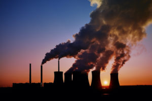 SMOKE BILLOWS OUT OF THE TOP OF SMOKESTACKS AT A POWER PLANT IN FRONT OF A SETTING SUN.