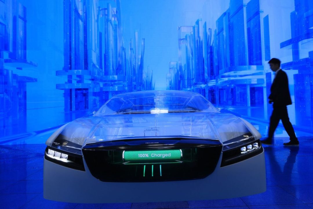 A CAR WITH EV TECHNOLOGIES READS "100% CHARGED" ON ITS FRONT BUMPER. THE CAR IS SITTING IN FRONT A BLUE SCREEN WITH A PERSON TO THE RIGHT OF IT.