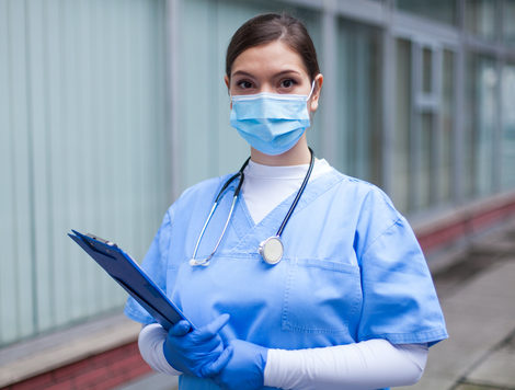 A NURSE IN BLUE SCRUBS AND A WHITE TURTLENECK WEARING A MASK LOOKS INTO THE CAMERA WHILE HOLDING A CLIPBOARD.