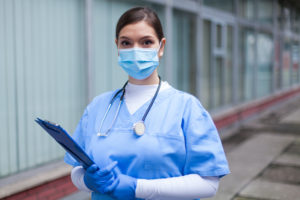 A NURSE IN BLUE SCRUBS AND A WHITE TURTLENECK WEARING A MASK LOOKS INTO THE CAMERA WHILE HOLDING A CLIPBOARD.