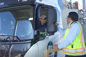 A TRUCK DRIVER LEANS THROUGH HIS CAB WINDOW TO GREET A SMILING PERSON IN A HI-VIS VEST