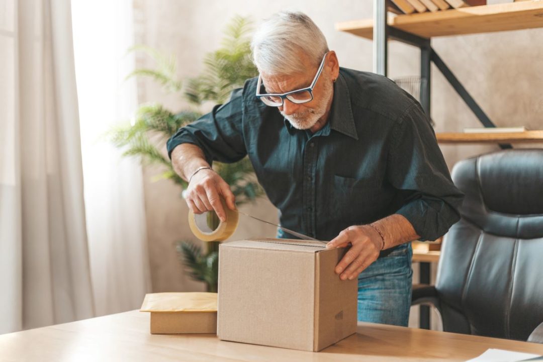 A MAN APPLIES PACKING TAPE TO A BROWN CARDBOARD BOX IN A LIVING ROOM OR OFFICE