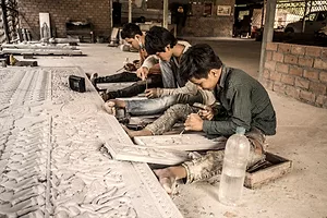 A ROW OF CHILDREN SITS CARVING WOODEN PANELS ON A DIRT FLOOR.