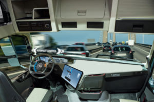 INTERIOR OF A SELF-DRIVING TRUCK. THE PHRASE "AUTOPILOT CONTROL" HOVERS ABOVE THE STEERING WHEEL. OUTSIDE THE FRONT WINDOW, SEVERAL CARS DRIVE ON A BRIDGE.