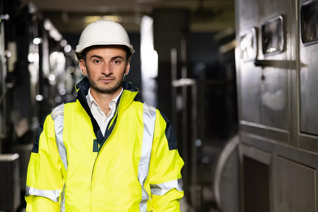 A FRONTLINE WORKER IN A YELLOW JACKET AND HARDHAT LOOKS INTO THE CAMERA.