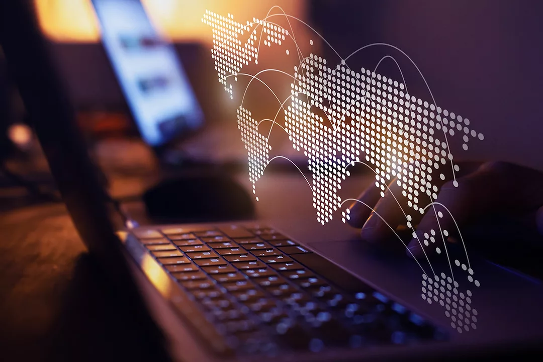 A DIGITAL MAP OF THE WORLD HOVERS ABOVE A LAPTOP'S KEYBOARD. CERTAIN PARTS OF THE MAP ARE CONNECTED BY CURVED LINES.