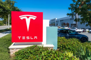 A TESLA SIGN SITS IN FRONT OF A PARKING LOT CONNECTED TO A TESLA DELIVERY CENTER.