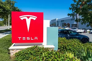A TESLA SIGN SITS IN FRONT OF A PARKING LOT CONNECTED TO A TESLA DELIVERY CENTER.