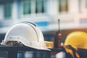 A WALKIE TALKIE SITS ON A TABLE BETWEEN YELLOW AND WHITE HARDHATS. THE YELLOW HARDHAT IS ON THE LEFT. THE WHITE HARDHAT IS ON THE RIGHT. 