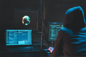 A PERSON IN A HOODIE CAN BE SEEN WORKING ON A COMPUTER WITH MULTIPLE MONITORS IN A DARK ROOM.