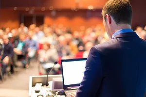 A SUIT-CLAD SPEAKER AT A PODIUM ADRESSES A BLURRED AUDIENCE