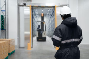 A WAREHOUSE WORKER LOOKS THROUGH A DOOR AT ANOTHER WORKER IN A COLD STORAGE FACILITY ON A FORKLIFT.