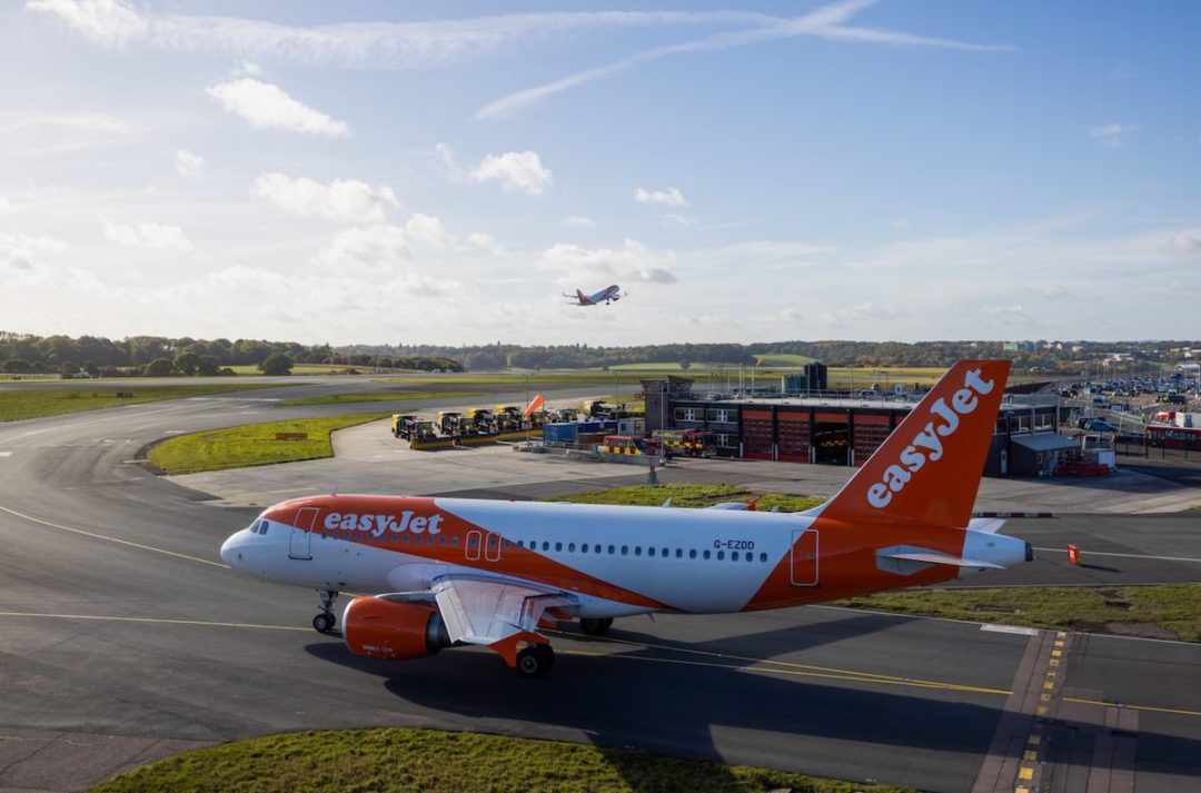 AN ORANGE AND WHITE EASYJET AIRPLANE CAN BE SEEN ON A RUNWAY. AN AIRPLANE IS FLYING IN THE BACKGROUND OVER THE RUNWAY.
