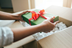 A GREEN AND RED WRAPPED CHRISTMAS GIFT IS BEING TAKEN OUT OF A CARDBOARD BOX WITH TWO HANDS.