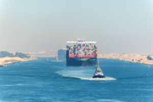 A LARGE CONTAINER SHIP TRAVELING ON THE SUEZ CANAL SURROUNDED BY TUGBOATS.
