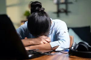 A YOUNG WOMAN RESTS HER HEAD IN HER FOLDED ARMS ON TOP OF AN OPEN LAPTOP
