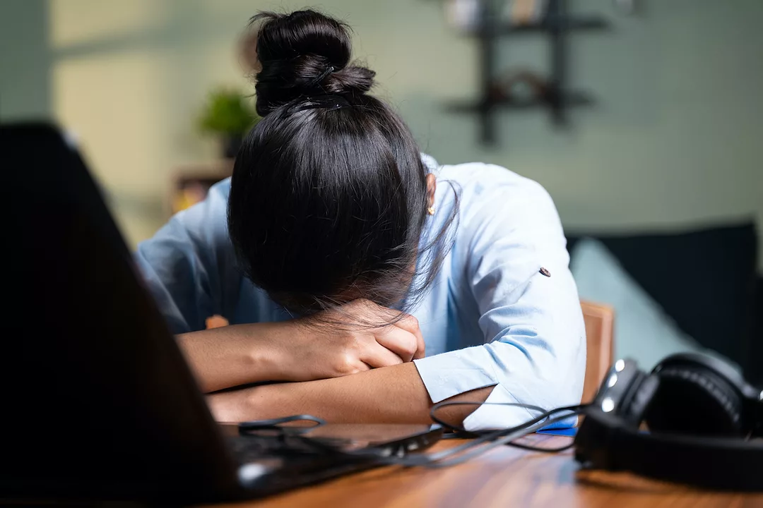 A YOUNG WOMAN RESTS HER HEAD IN HER FOLDED ARMS ON TOP OF AN OPEN LAPTOP