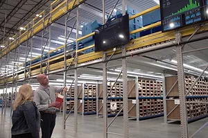 TWO WORKERS IN A WAREHOUSE STAND BENEATH A GIANT SCREEN WITH GRAPHS ON IT