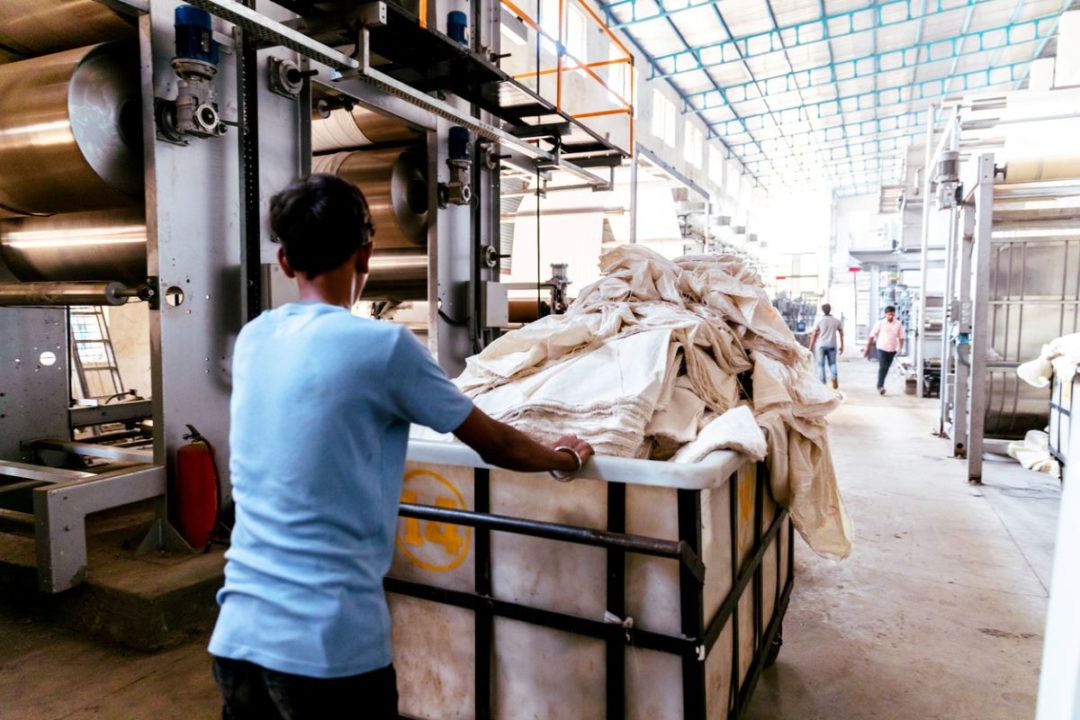 A WORKER PUSHES AN INDUSTRIAL BIN FULL OF TEXTILES THROUGH A WAREHOUSE AISLE