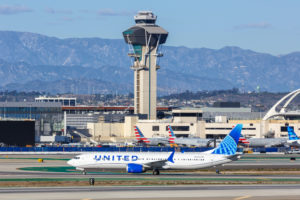 A UNITED AIRLINES BRANDED BOEING 737 MAX 9 JET SITS ON A RUNWAY IN FRONT OF AN AIRPORT AND CONTROL TOWER.