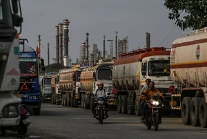 MOTORCYCLISTS DRIVE BY OIL TANKER TRUCKS OUTSIDE OF A REFINERY.