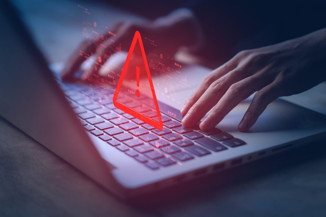 A DIGITAL RENDERING OF A RED TRIANGLE WITH AN EXCLAMATION POINT INSIDE OF IT HOVERS ABOVE A PERSON'S HANDS ON A LAPTOP.