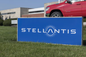 A STELLANTIS SIGN SITS ON THE GRASS IN FRONT OF A RED CAR.