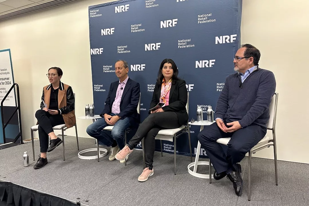 FOUR PEOPLE SIT ON CHAIRS ON A STAGE, FRAMED BY AN NRF BACKDROP