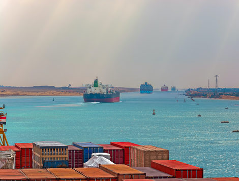 LARGE CONTAINER SHIPS TRAVEL ON THE SUEZ CANAL WITH CONTAINERS IN THE FOREGROUND OF THE PHOTO.