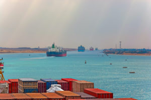 LARGE CONTAINER SHIPS TRAVEL ON THE SUEZ CANAL WITH CONTAINERS IN THE FOREGROUND OF THE PHOTO.