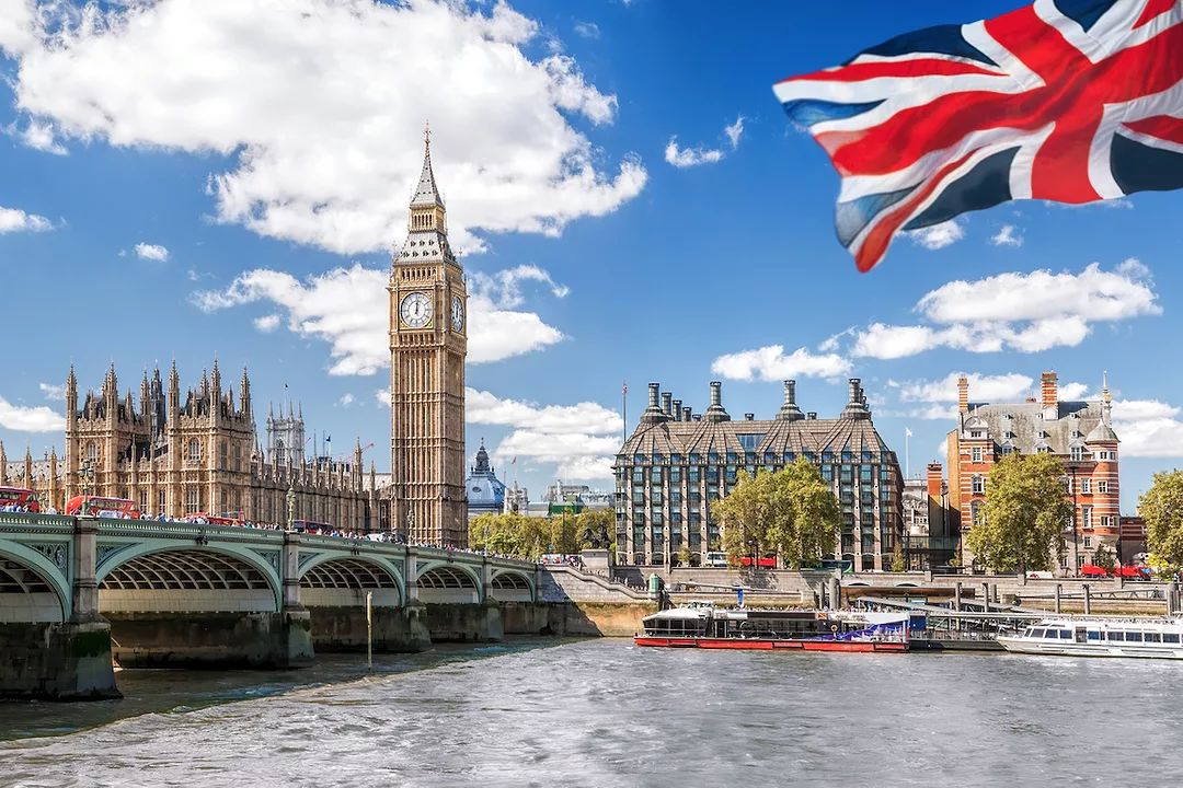THE BRITISH FLAG WAVING IN FRON OF BIG BEN AND THE BRIDGE OVER THAMES UNDER A BLUE SKY.