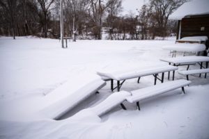 THREE PICNIC TABLES AND A FIELD ARE COVERED IN SNOW.