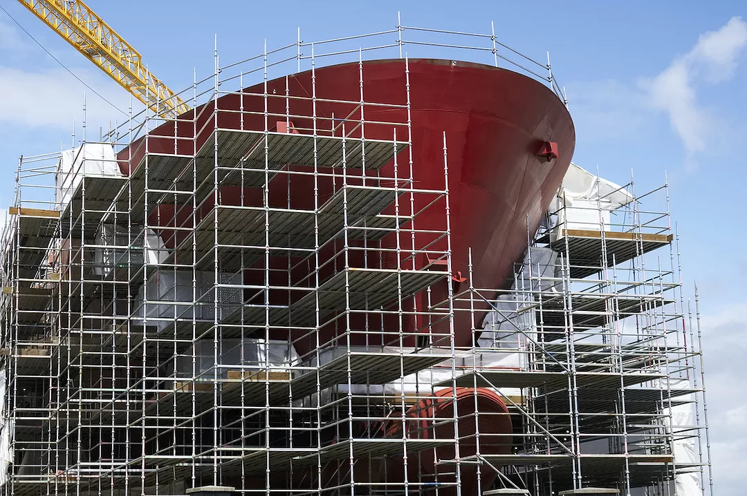 SCAFFOLDING AND A CRANE SURROUNDS A LARGE SHIP BEING BUILT.