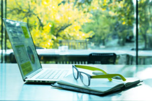 A GREEN AND BLUE PAIR OF GLASSES SIT ON TOP OF A NOTEBOOK NEAR AN OPEN LAPTOP WITH GREEN GRAPHS ON IT. MANY LUSH, GREEN TREES CAN BE SEEN THROUGH A LARGE WINDOW IN THE BACKGROUND.