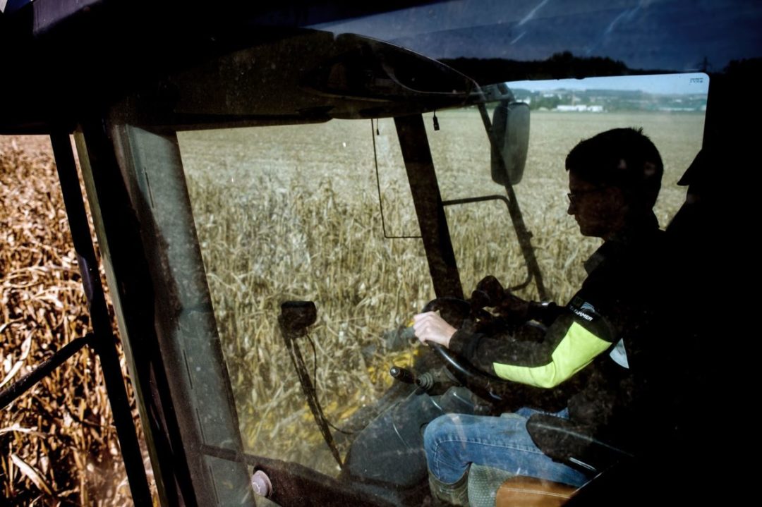A FARM WORKER SITS IN THE CAB OF AN AGRICULTURAL VEHICLE SURROUNDED BY FIELDS OF GRAIN