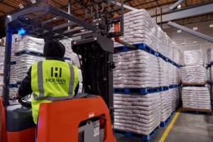 A WORKER SEEN FROM BEHIND OPERATES A FORKLIFT TRUCK IN A WAREHOUSE WITH WHITE BAGS STACKED HIGH