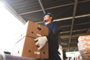 GROUND VIEW OF A PERSON IN A CONSTRUCTION HELMET CARRYING TWO BOXES STACKED ON TOP OF EACH OTHER.