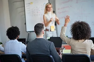 A PERSON GIVING A SPEECH IN FRONT OF A WHITEBOARD TO THREE PEOPLE SEATED AT A TABLE. THE PERSON TO THE RIGHT HAD THEIR HAND RAISED.